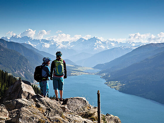 2 Personen mit Helm stehen auf einem Felsen und blicken über einen See