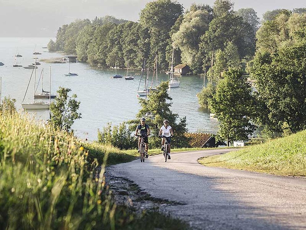 2 Radfahrer auf einer Straße, im Hintergrund der Attersee mit Segelbooten