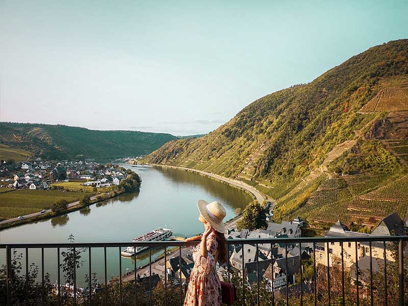 Eine Dame mit Hut steht auf einer Brücke über die Mosel, im Hintergrund der Ort Beilstein und Weinberge.