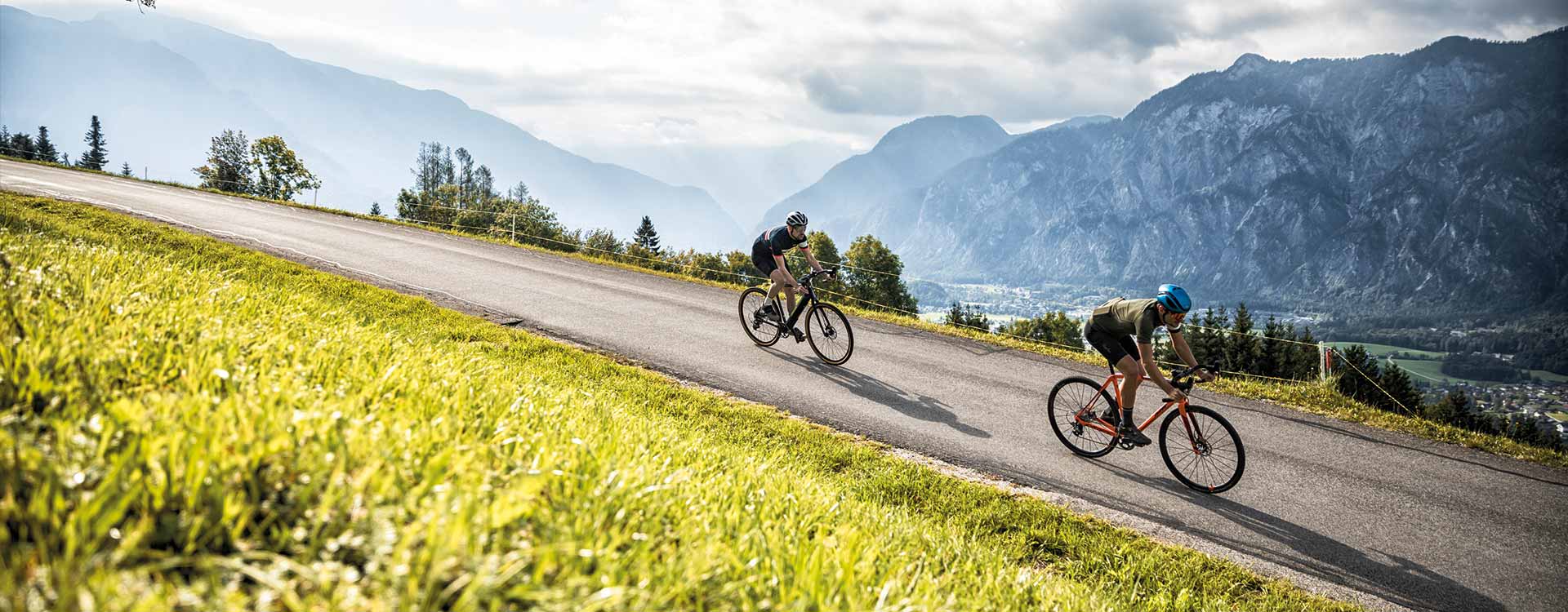 2 Gravelbiker auf einer Straße, im Hintergrund Bergpanorama