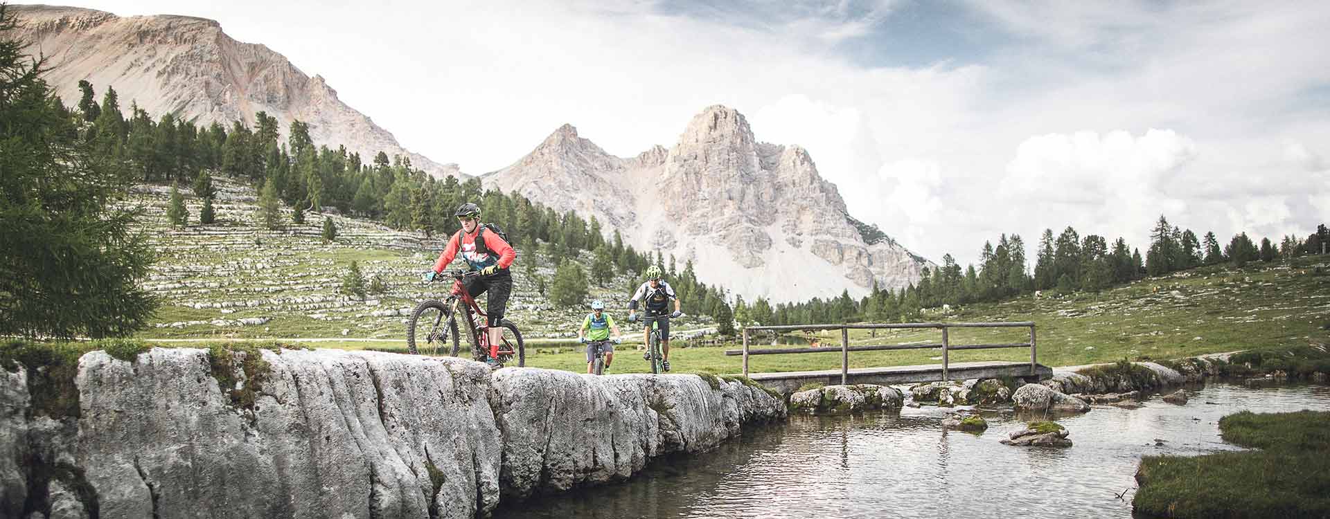3 Biker fahren entlang eines Gebirgsbaches, im Hintergrund Bergpanorama