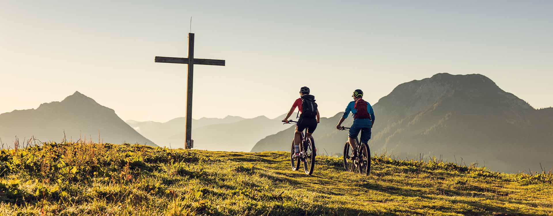 2 Biker fahren auf das Gipfelkreuz zu. Im Hintergrund Bergpanorama.