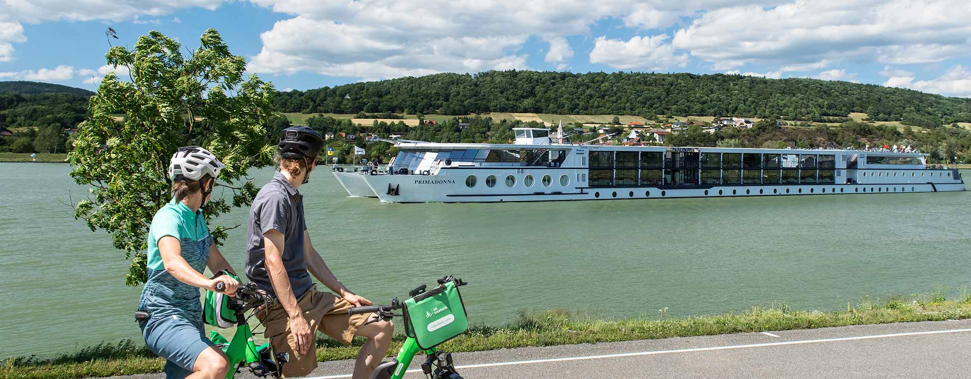 2 Radfahrer am Ufer, im Hintergrund die MS Primadonna auf der Donau