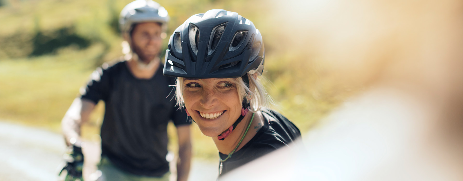 eine Dame mit Radhelm lächelt freundlich in die Kamera, im Hintergrund ein weiterer Radfahrer, der verschwommen ist.