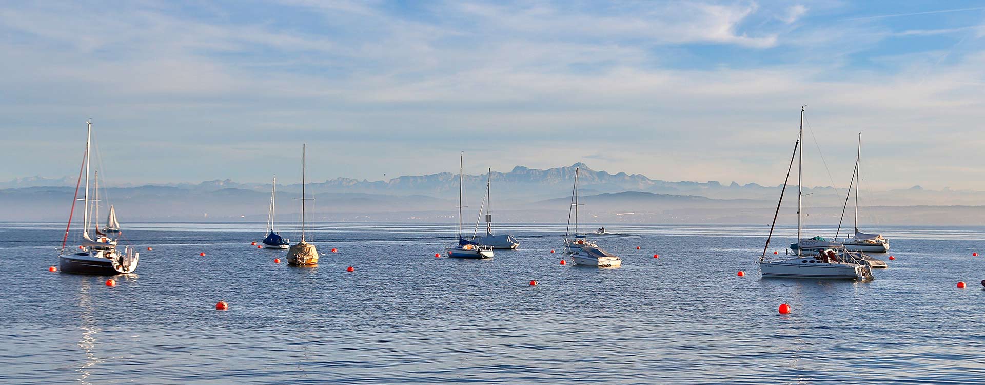 Panoramabild über den Bodensee mit Segelschiffen, im Hintergrund erkennt man einen Gebirgszug