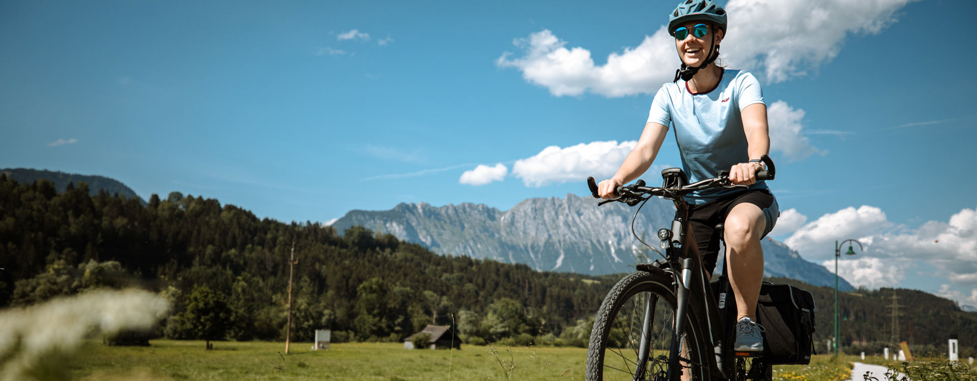 eine Dame am Fahrrad radelt am flachen Radweg, im Hintergrund Bergpanorama