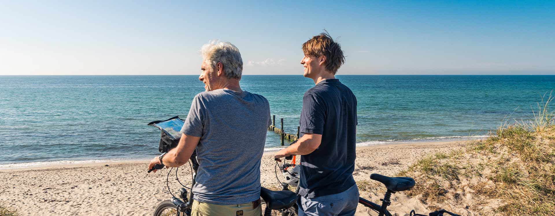 2 Männer schieben ihre Fahrräder in Richtung Sandstrand am Ostseeufer.