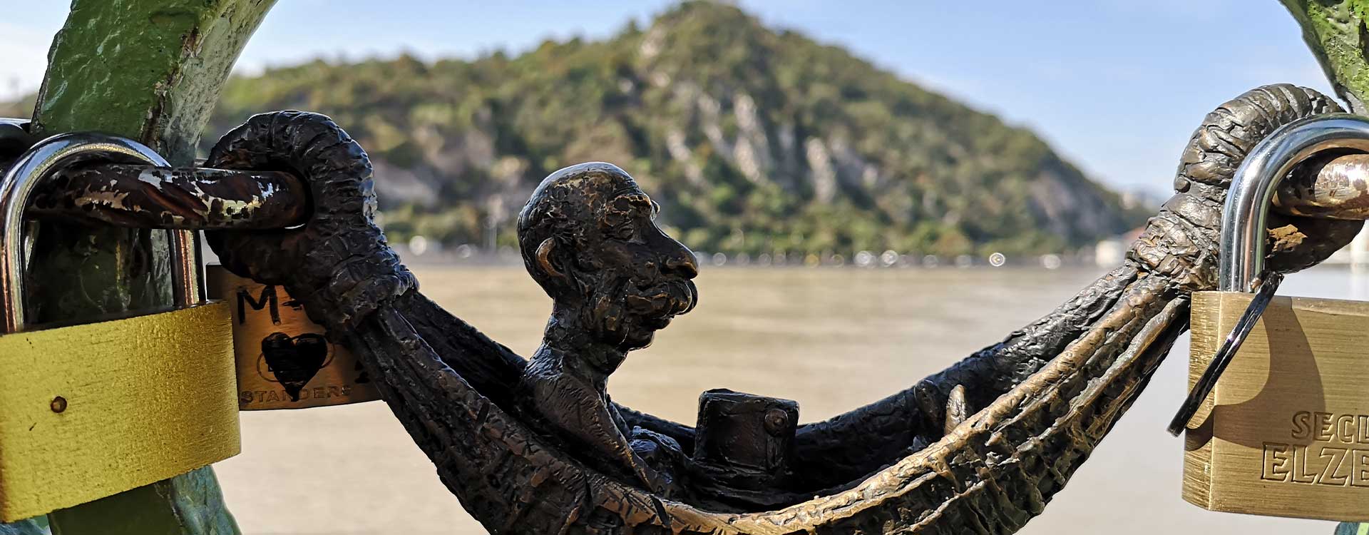 Eine Figur auf einem Brückengeländer, daneben Liebesschlösser. Im Hintergrund die Donau bei Budapest.