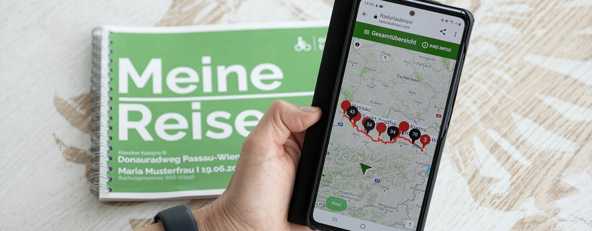 eine Hand hält ein Smartphone, auf dem die digitale Anwendung mit Tagesetappe zu sehen ist. Im Hintergrund das gedruckte Routenbuch.